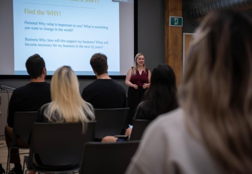A presenter stands in front of a screen displaying text, facing an audience seated in chairs.