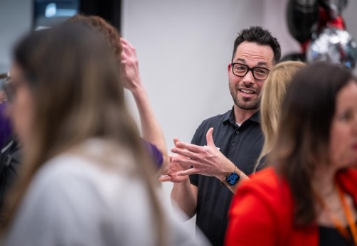 A person with glasses gestures with open hands while surrounded by others.