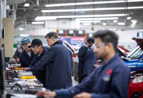 At the Centre for Applied Transportation Technology at Fanshawe College, several people in navy blue work shirts and safety glasses are gathered around a workbench with a computer, tools, and papers, while other people work on vehicles with open hoods in the background.
