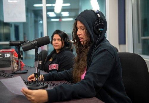 Two women in a recording studio. One speaks into a microphone, wearing headphones and a hoodie, while the other listens attentively. The environment is bright and focused.