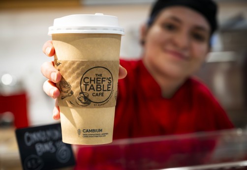A hand holds a paper coffee cup with a white lid, featuring the Chef's Table Cafe logo, at the London Downtown Campus of Fanshawe College.