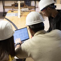 Two students with Construction Project Management instructor, reviewing plans on laptop at job site