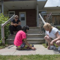 Three people sitting outside; two drawing on the concrete with chalk while another watches