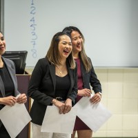 Three female students smiling and laughing while presenting an operations project in front of their class.