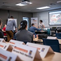 A classroom setting with tables and blue chairs, a projector screen displaying a video call, and a whiteboard with notes.