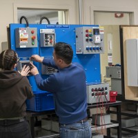 Two people stand in front of an Industrial Control Trainer, which is a blue board with various electrical components and warning signs attached.