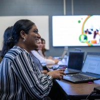 At Fanshawe College's London Campus, a business class is in session, with a person in a striped shirt smiling while holding a phone, surrounded by laptops on a table, and a presentation