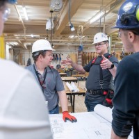 In a Fanshawe College workshop, individuals wearing hard hats and safety glasses examine a large blueprint spread across a table.