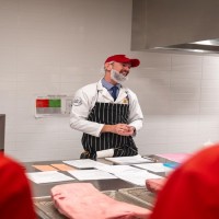 At the London Downtown Campus, in a room with white tiled walls and stainless steel counters, a person in a white lab coat, red baseball cap, and black and white striped apron stands behind a counter with papers and a large cut of meat, while two people in red jackets stand in the foreground.