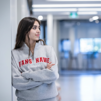 A person wearing a grey sweater with red lettering leans against a white wall in a hallway.