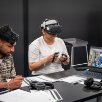 One person writes on documents while another wears a virtual reality headset and holds handheld controllers at a table featuring a laptop displaying a virtual environment.