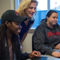 Photo of a women looking at a computer screen while two students are sitting down looking at the screen as well