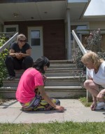 Three people sitting outside; two drawing on the concrete with chalk while another watches