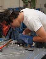 At the Centre for Applied Transportation Technology at Fanshawe College, a person wearing a flame-designed welding mask and blue gloves is bent over a gray metal car part, possibly a door, using a tool that is creating sparks. A red hammer with a wooden handle rests on the metal surface. Behind the person is a red gas tank with gauges and a chain, and various pieces of machinery.