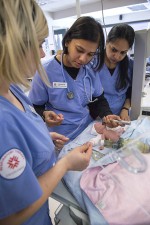 In the School of Health Sciences at Fanshawe College's London Campus, three people in blue scrubs are gathered around a baby mannequin on a light blue blanket, with medical equipment nearby.